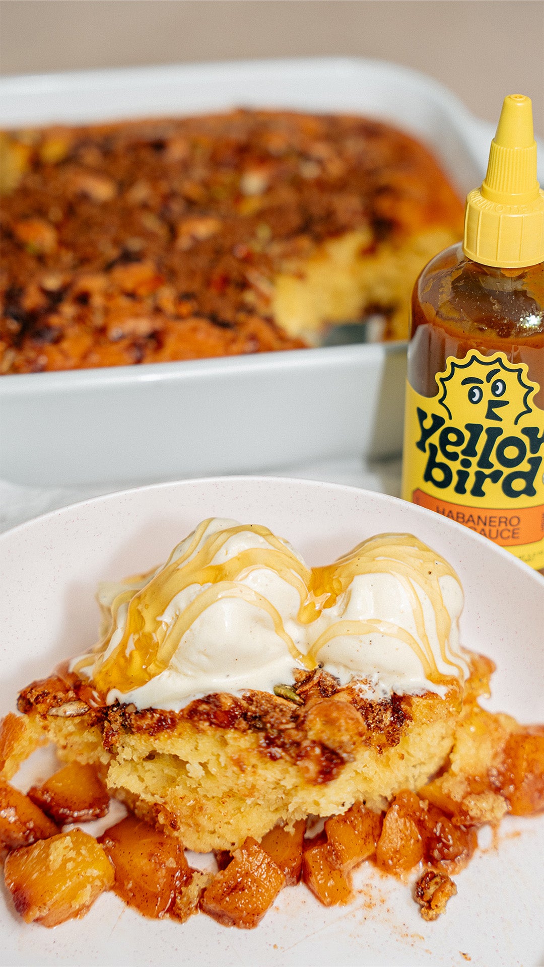 A piece of cake topped with ice cream and honey in the foreground, with a bottle of Yellowbird Habanero Hot Sauce and the rest of the cake in the background.