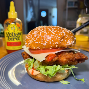 A spicy chicken sandwich on a toasted sesame brioche bun on a plate. There is a bottle of Yellowbird Jalapeño Hot Sauce in the background.
