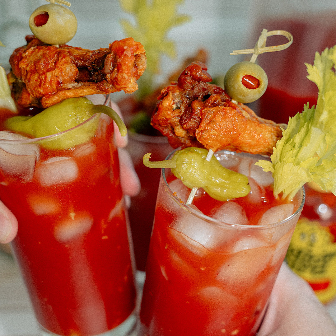 2 glasses of Bloody Mary doing "cheers", each has a skewer with a chicken wing, a peperoncino, and a green olive ,