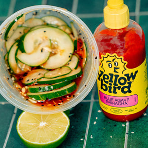 A deli container containing a marinated cucumber salad turned on its side, next to a bottle of Yellowbird Blue Agave Sriracha