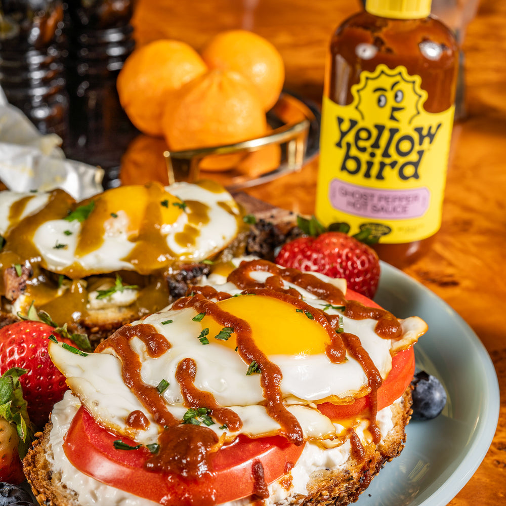 Yellowbird Organic Ghost Pepper Hot Sauce drizzled onto fried egg, tomato, and cottage cheese toast on a wooden table.