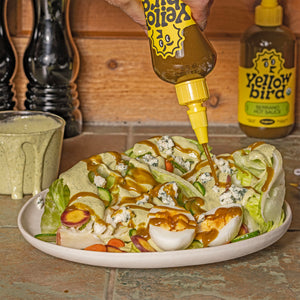 Crisp iceberg lettuce wedges topped with creamy Serrano herb dressing, fresh cucumbers, radishes, carrots, blue cheese crumbles, hardboiled eggs, and a drizzle of Yellowbird Organic Serrano Hot Sauce.
