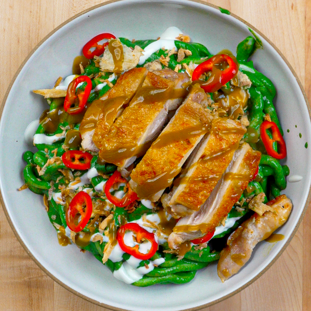 Overhead shot of udon noodles in a white bowl, coated in a green Serrano sauce, topped with pan-seared chicken, shallots, red chili peppers, sour cream, and an extra drizzle of Yellowbird Serrano Hot Sauce.