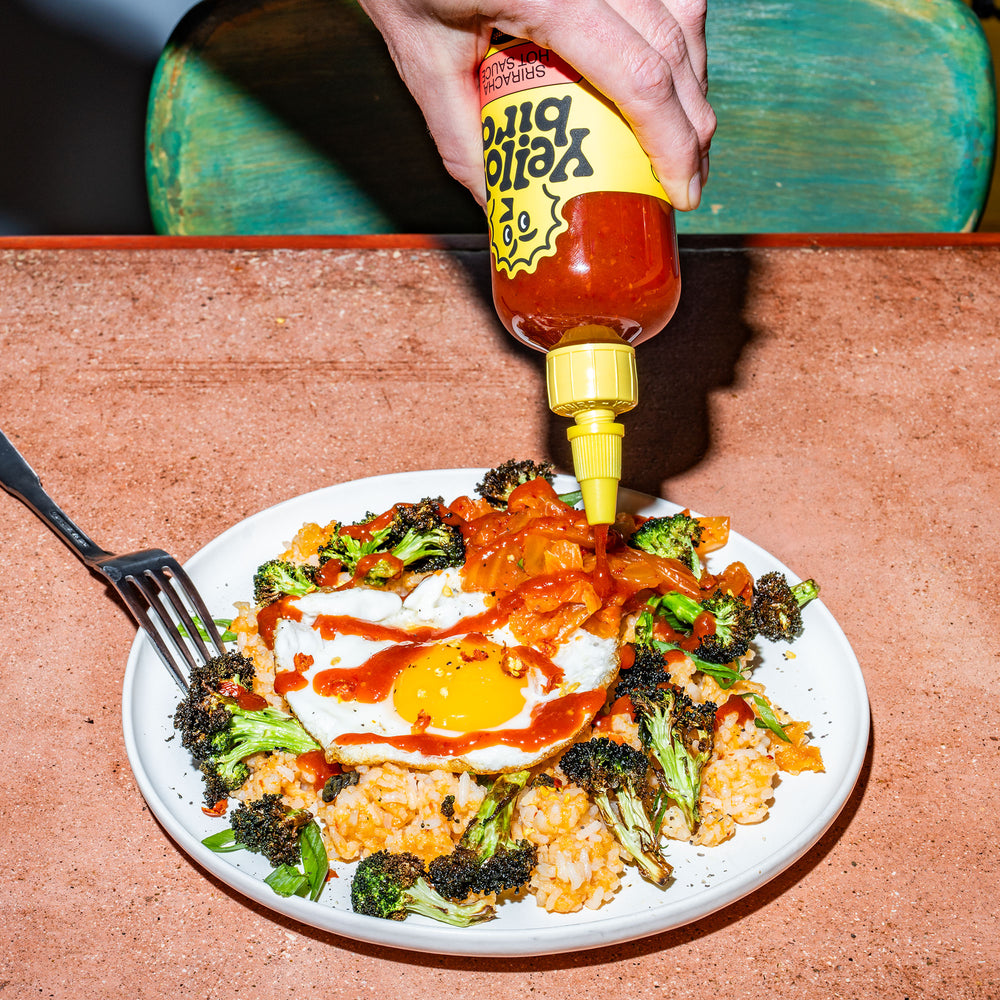 Person pouring Yellowbird Organic Sriracha hot sauce over a plate of fried rice with broccoli, a fried egg, and kimchi.